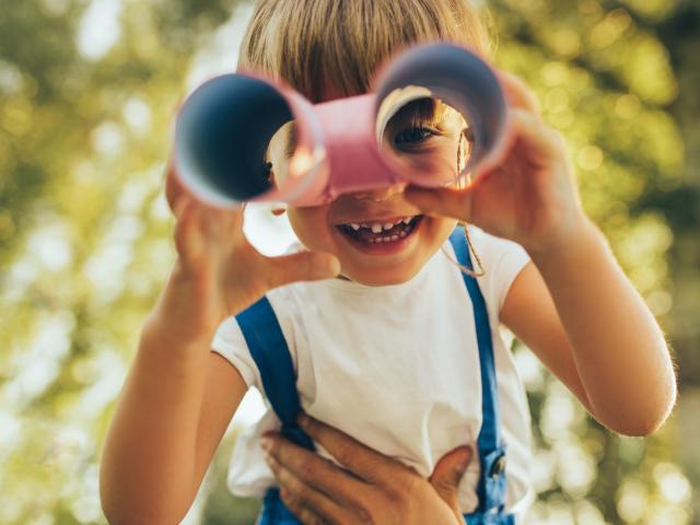 Closeup image of cute little boy playing with a binoculars searching for an imagination or exploration in summer day in park. Happy child playing pretend safari game outdoors in the forest. Childhood