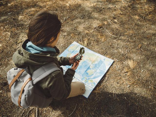 Traveler young woman with backpack searching direction with a compass on background of map in the forest. Focus on compass