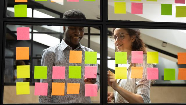 Multiracial colleagues developing business ideas use post-it notes view through glass, mates read written tasks shown on stickers attached on wall, effective way do work don't forget anything concept