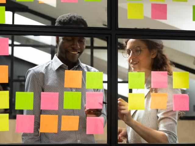 Multiracial colleagues developing business ideas use post-it notes view through glass, mates read written tasks shown on stickers attached on wall, effective way do work don't forget anything concept