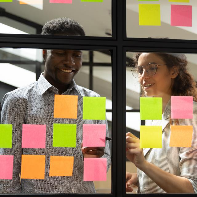 Multiracial colleagues developing business ideas use post-it notes view through glass, mates read written tasks shown on stickers attached on wall, effective way do work don't forget anything concept